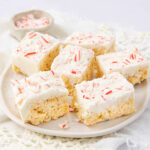A plate of rice crispy treats topped with a layer of white frosting and sprinkled with crushed peppermint candy pieces, arranged on a white tablecloth.