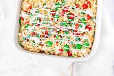 A square dish of Rice Krispies treats topped with red and green M&M candies, colorful sprinkles, and white chocolate drizzle, sitting on a white surface.