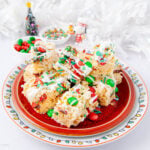 A festive plate of rice krispie treats topped with colorful holiday sprinkles, red and green candies, and drizzled white chocolate. Christmas decorations and candy bowls are in the background.