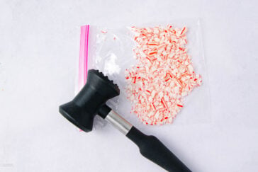 A black mallet rests next to a ziplock bag containing crushed peppermint candy pieces on a white surface.