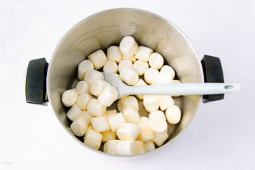 A metal pot filled with large white marshmallows and a white spatula resting inside, seen from above on a white surface.