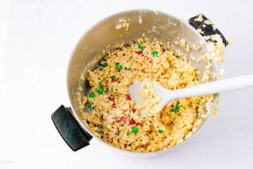 A mixing bowl filled with Rice Krispies cereal, melted marshmallow mixture, and colorful candy-coated chocolates, with a white spatula resting inside the bowl.