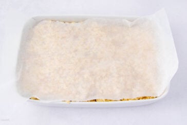 A white baking dish filled with food is covered by a sheet of parchment paper, partially tucked into the sides. The dish is set on a white surface.