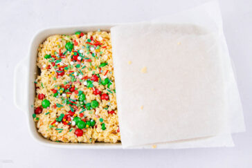 A baking dish with holiday rice crispy treats topped with red, green, and white sprinkles and candies, partially covered by a sheet of parchment paper.