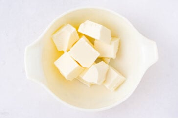 A white mixing bowl containing several cubes of butter on a white surface.
