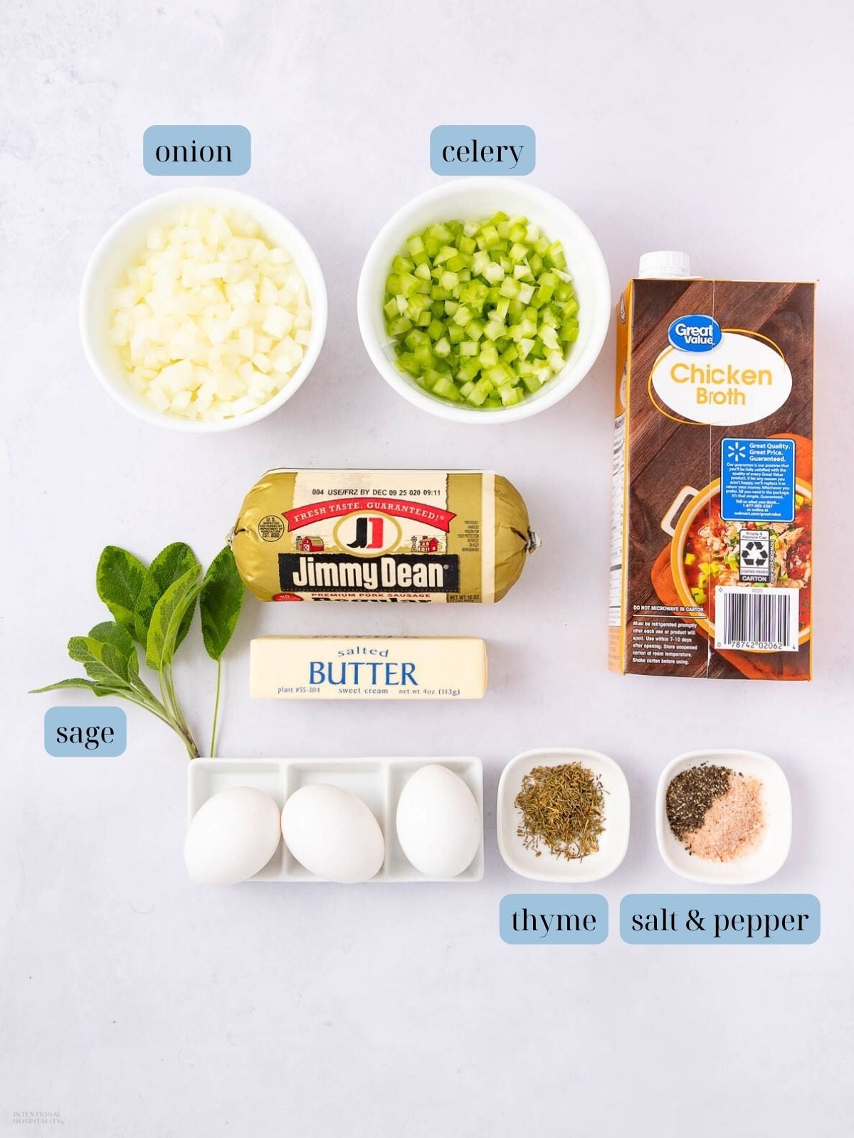 Flat lay of stuffing ingredients: diced onion, chopped celery, chicken broth carton, breakfast sausage roll, fresh sage leaves, stick of butter, three eggs, dried thyme, salt, and pepper in small dishes.