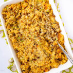 A white baking dish filled with golden-brown cornbread stuffing, mixed with herbs and vegetables, with a silver serving spoon resting inside. The dish sits on a patterned cloth.