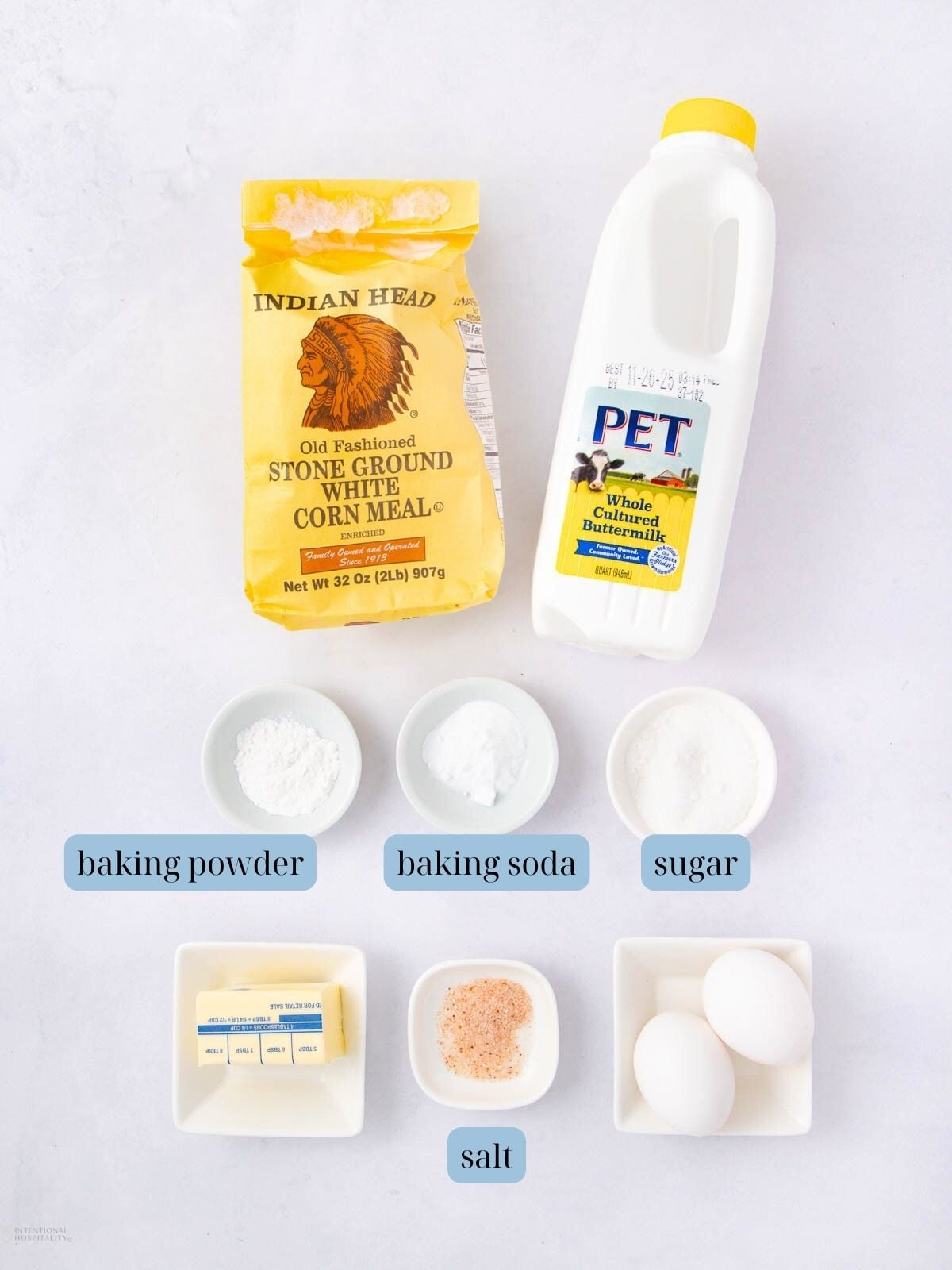 A bag of stone ground white cornmeal, a bottle of buttermilk, bowls of baking powder, baking soda, sugar, salt, two eggs, and a dish of butter are arranged on a white surface with labels for each ingredient.