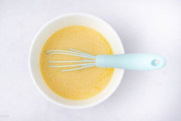 A light blue whisk rests in a white bowl filled with a yellow batter or mixture, set against a plain white background.