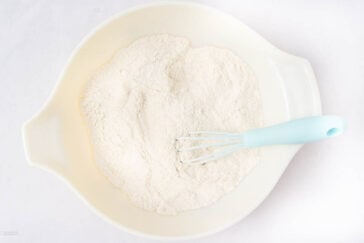 A white mixing bowl filled with flour and a light blue whisk resting inside, set on a white surface.