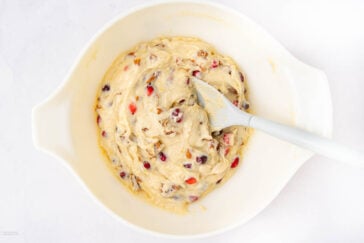 A white mixing bowl filled with thick batter containing red and brown dried fruits, with a white spatula resting inside. The bowl is placed on a white surface.