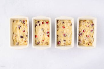 Four small white baking dishes, each filled with raw batter containing visible pieces of cranberries and nuts, are arranged in a row on a white surface.