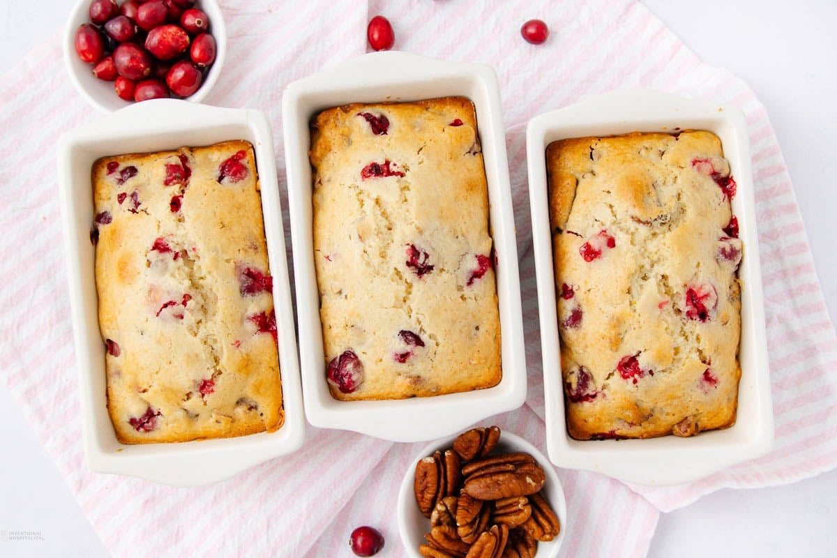 Three small white loaf pans filled with golden brown cranberry bread, surrounded by fresh cranberries and pecans on a pink-striped cloth.