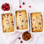 Three small loaves of cranberry bread in white loaf pans sit on a light pink striped cloth, with a bowl of fresh cranberries, a bowl of pecans, and a few scattered cranberries and pecans nearby.