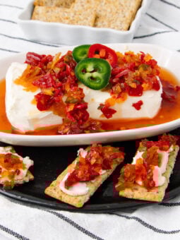 A block of cream cheese topped with red pepper jelly and sliced jalapeños on a white dish, served with crackers, some topped with the same spread, on a black plate with a striped cloth underneath.