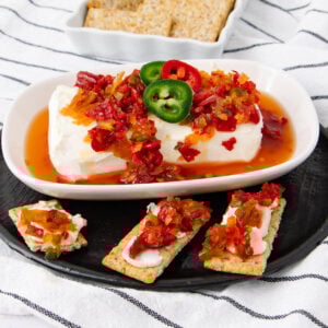 A block of cream cheese topped with red pepper jelly and sliced jalapeños on a white dish, served with crackers, some topped with the same spread, on a black plate with a striped cloth underneath.