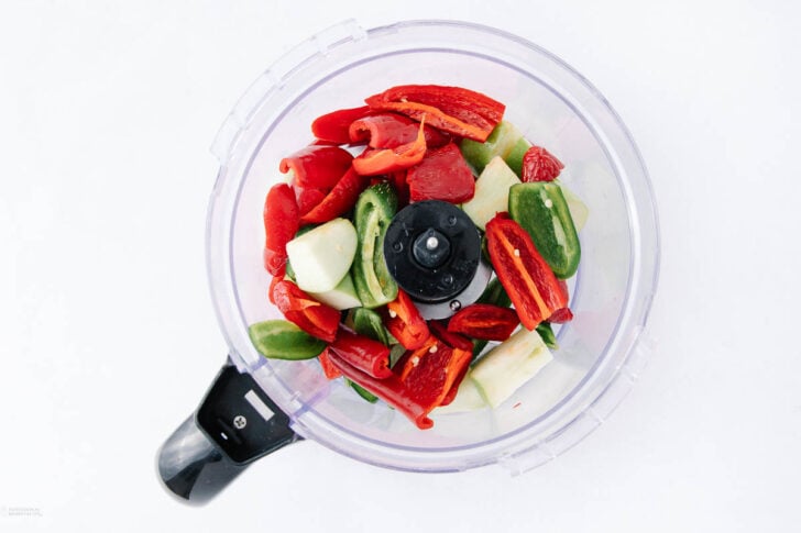 A food processor bowl containing chopped red and green peppers and pieces of white onion, viewed from above on a white background.