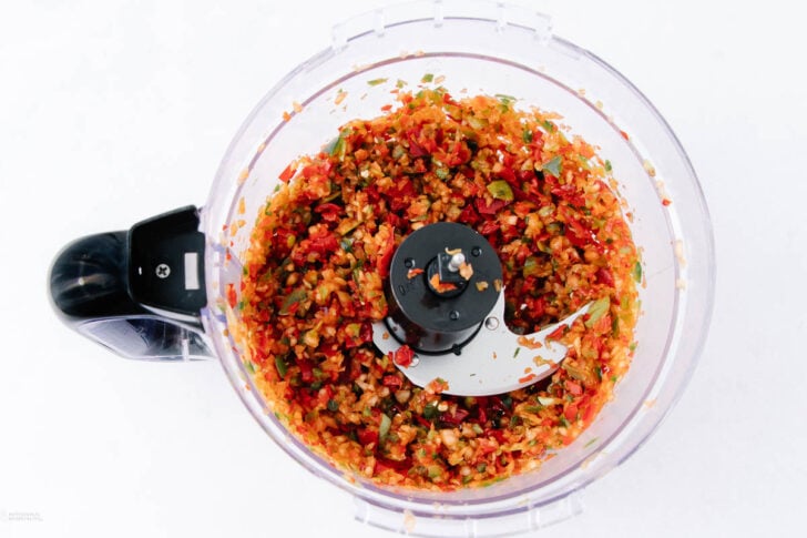 A food processor filled with finely chopped red, green, and orange vegetables, viewed from above on a white background.