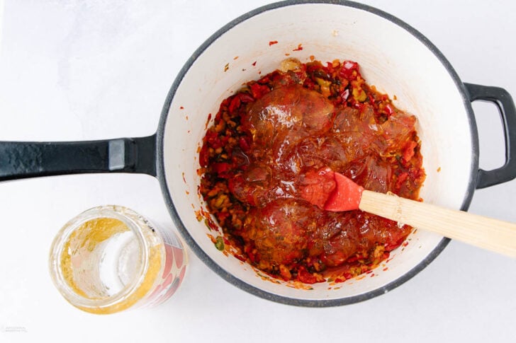 A pot with chopped vegetables and tomato sauce being stirred by a red spatula, next to an empty glass jar on a white surface.