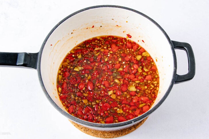 A black and white pot filled with chunky chili oil containing chopped red chilies, garlic, and oil, placed on a round cork trivet against a white background.