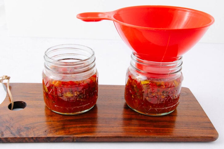Two glass jars filled with red sauce sit on a wooden board. One jar is open, the other has a wide red funnel on top, ready for filling. The background is white.
