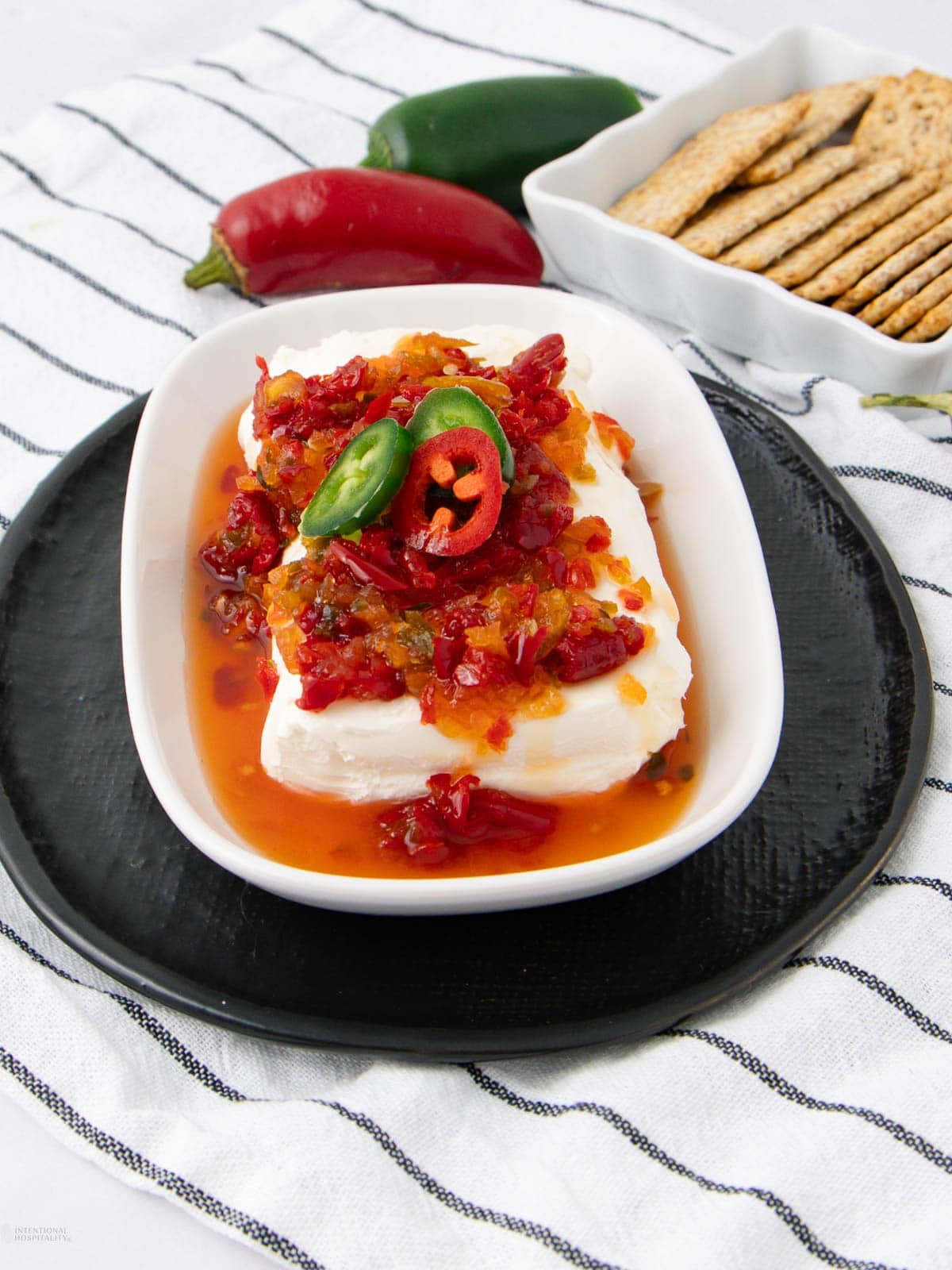 A block of cream cheese topped with red and yellow pepper jelly, garnished with sliced jalapeños, served on a white dish with crackers and whole chili peppers in the background.