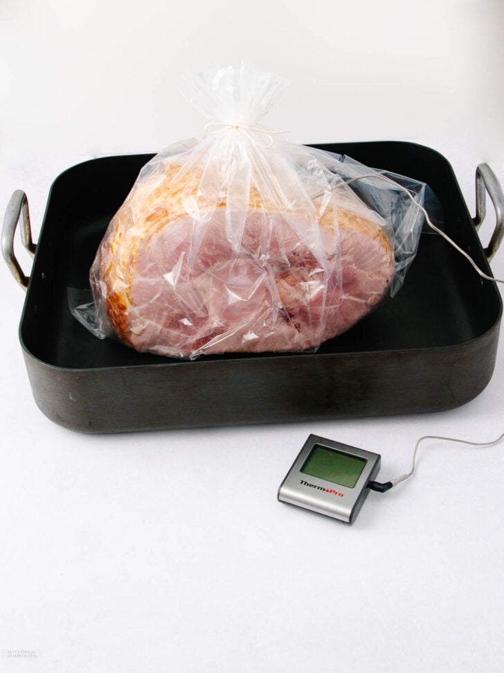 A large ham wrapped in a plastic roasting bag sits in a black roasting pan. A meat thermometer probe is inserted into the ham, with the digital display resting on the white surface next to the pan.