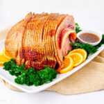 A sliced glazed ham on a white platter, garnished with fresh parsley and orange slices, with a small bowl of sauce on the side. The platter sits on a beige cloth.