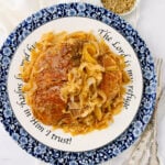 A plate with two pieces of browned meat smothered in caramelized onions and gravy, served on a decorative blue and white plate with the text, “The Lord is my refuge, in Him I trust!” around the rim.