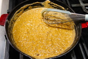 A close-up of a bubbling brown sauce being whisked in a red cast iron skillet on a stove, with the whisk resting in the thick mixture.
