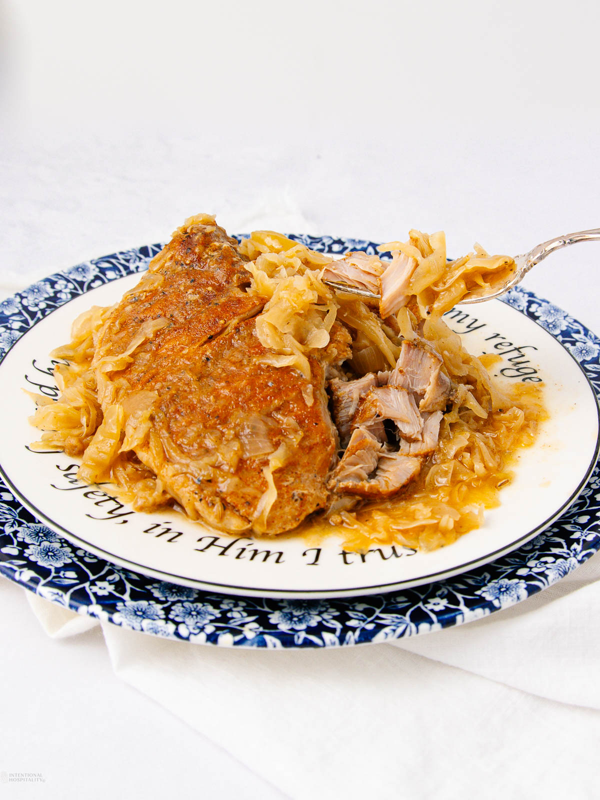 A plate featuring a serving of braised pork and sauerkraut with shredded pieces, set on a decorative plate with a blue and white border, on a white tablecloth.