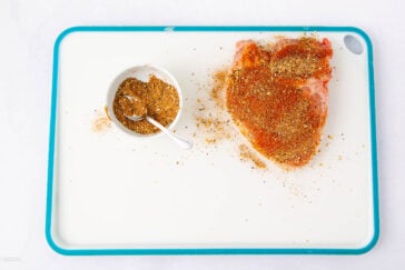 A white cutting board with a blue border holds a raw, seasoned piece of meat and a small white bowl of seasoning with a spoon. Some seasoning is scattered on the board.
