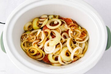 A white slow cooker filled with raw seasoned meat covered with a generous layer of sliced onions, ready for cooking.