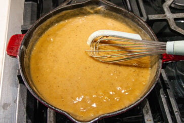 A skillet filled with thick brown gravy being whisked on a stovetop, with a metal whisk and spatula in the pan.