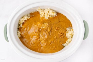 A white slow cooker filled with shredded sauerkraut around the edges and covered with a thick, brownish-orange sauce in the center.
