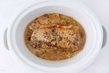 A cooked pork roast with a seasoned crust sits in broth and cabbage inside a white slow cooker, viewed from above.