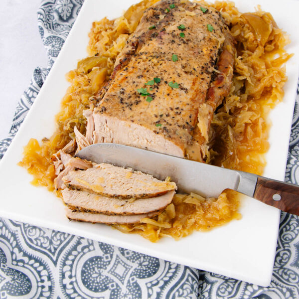 A sliced pork roast with herbs sits on a white platter, surrounded by cooked cabbage and onions. A knife rests beside the partially sliced meat. The platter is on a patterned cloth.