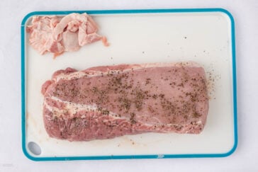 A large, raw pork loin seasoned with herbs sits on a white cutting board with a blue border. Trimmed fat pieces are placed to the side of the meat.