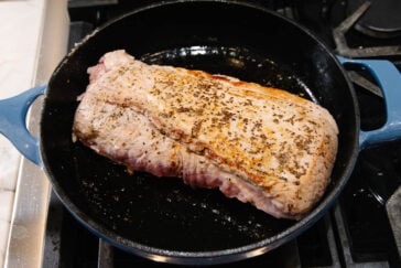 A large piece of seasoned pork is searing in a blue cast iron skillet on a stovetop, with a golden-brown crust forming on the surface.