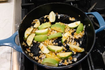 A blue cast iron skillet on a stovetop contains chopped onions and green apple slices being sautéed. The onions appear golden and the apples are partially cooked.