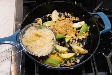 A blue skillet on a stove contains chopped onions, apple slices, and brown sugar, with a mesh strainer holding sauerkraut positioned over the pan, ready to be added.
