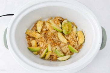 A white slow cooker containing sliced green apples, sauerkraut, and brown lentils or grains, partially mixed together on a white background.