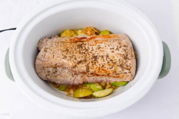 A browned pork roast seasoned with herbs sits on top of sliced green apples and onions inside a white slow cooker.