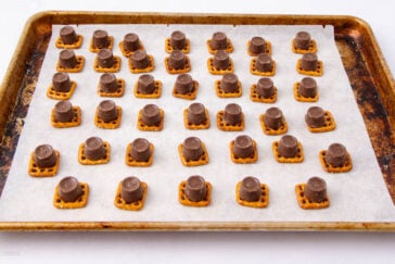 A baking sheet lined with parchment paper holds five rows of square pretzels, each topped with a chocolate-covered caramel candy, arranged in a neat grid pattern.