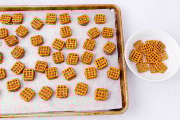 Square pretzel pieces are scattered on a parchment-lined baking sheet, with additional pretzels in a white bowl nearby, all set on a white surface.