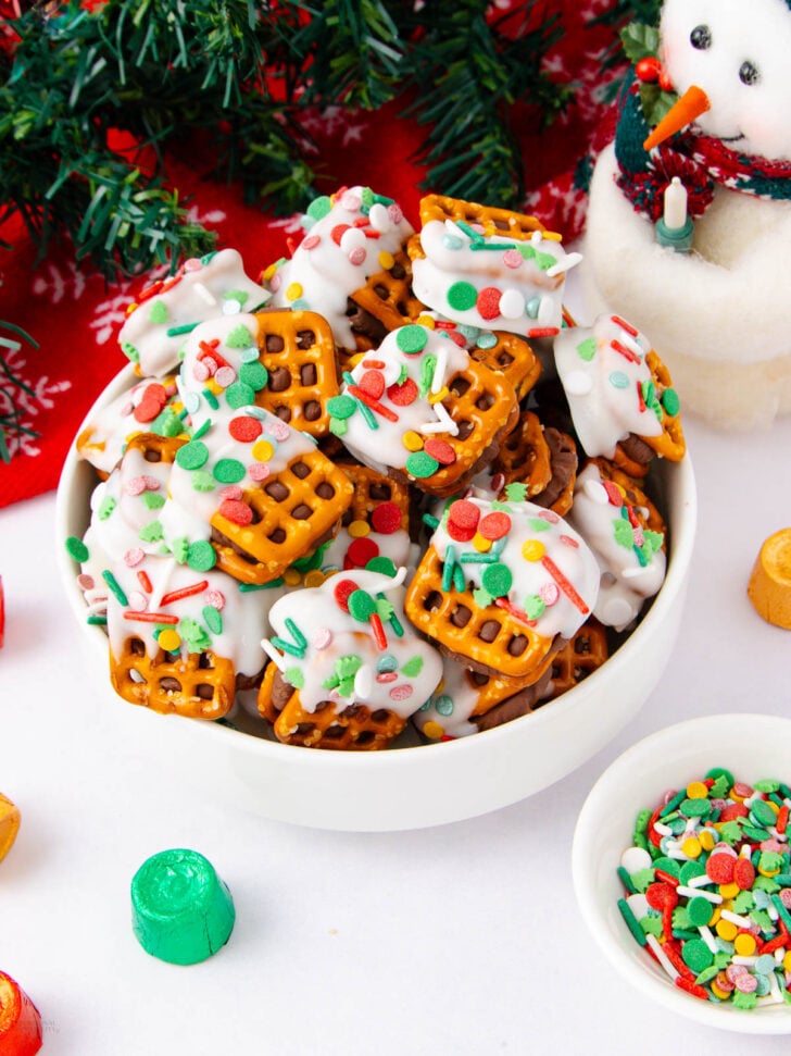 A bowl filled with pretzels dipped in white chocolate and decorated with colorful holiday sprinkles, surrounded by festive decorations, a snowman figurine, and a small bowl of sprinkles.