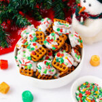 A white bowl filled with pretzels dipped in white icing and topped with red and green holiday sprinkles, sitting on a white surface near a snowman decoration, candies, and a Christmas tree.