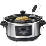 A stainless steel Hamilton Beach slow cooker with a digital display showing 180°F, a glass lid, a meat thermometer probe inserted, and food cooking inside, including onions and meat.