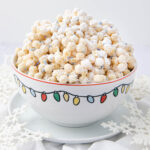 A festive bowl decorated with colorful holiday lights is filled with powdered sugar-coated popcorn and sprinkles, set on a white surface with snowflake-shaped decorations underneath.