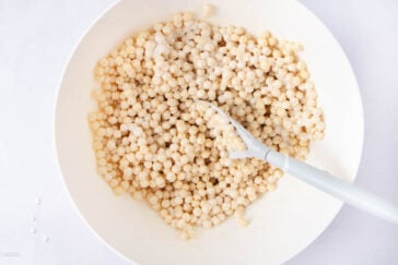 A white bowl filled with small, round, pale tapioca pearls coated in a creamy liquid. A white spoon is resting inside the bowl, mixing the pearls.